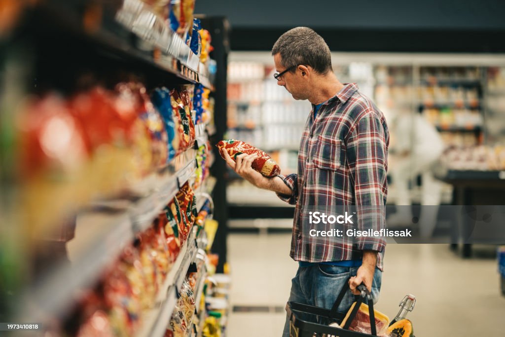 Man reading a label while grocery shopping in a supermarket aisle Man standing in a supermarket aisle and reading the label on a bag of pasta while grocery shopping Supermarket Stock Photo