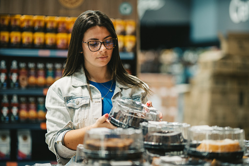 Young woman standing in a supermarket aisle and reading the label on a cake package while grocery shopping