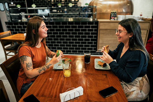 Side view of two women friends having fun talking and eating food at their table in restaurant