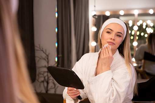 An exquisite image of a young makeup artist in her twenties, draped in a white bathrobe, gracefully cleansing her face in the soft glow of a well-lit mirror. The beige-toned room sets a warm ambiance, highlighting the dedication to skincare within the realm of makeup artistry. Perfect for capturing the serene and elegant side of beauty rituals.