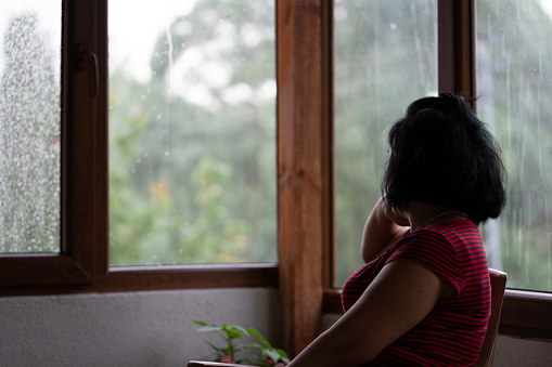 Candid photo of mature woman sitting on chair and looking through window. It is raining in outdoor. Shot indoor with a full frame mirrorless camera.