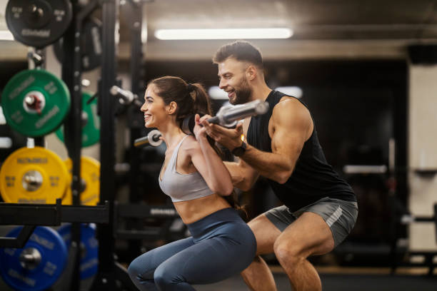 istock A fit woman is squatting with bar in a gym while her trainer helping her. 1973487171