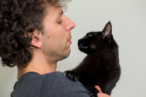 A young man with curly hair with anxiety is holding a cute pet black cat close and they are looking each other in the eye with love and affection.