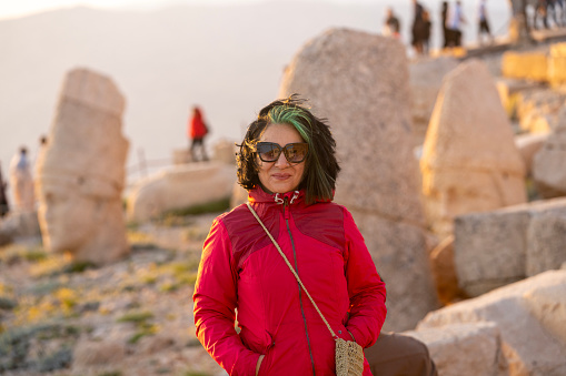 Photo of mature woman wearing a red coat and sunglasses standing next to monuomental rock statues on Nemrud Dagh, Adiyaman, Turkey. Statues are seen on the background. Shot under daylight during sunset with a ful lframe mirrorless camera.