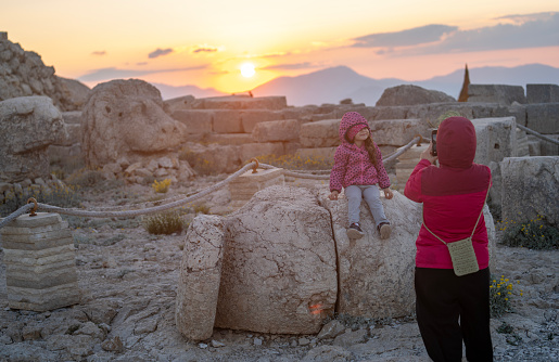 Candid photo of mother photographing 5 years old daughter in ancient ruins of Nemrut Dagi with large statues of ancient gods. Shot in outdoor with a full frame mirrorless camera.
