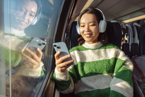 Photo of a young Asian woman sitting on the seat near the window on the train. She is smiling and using wireless technology