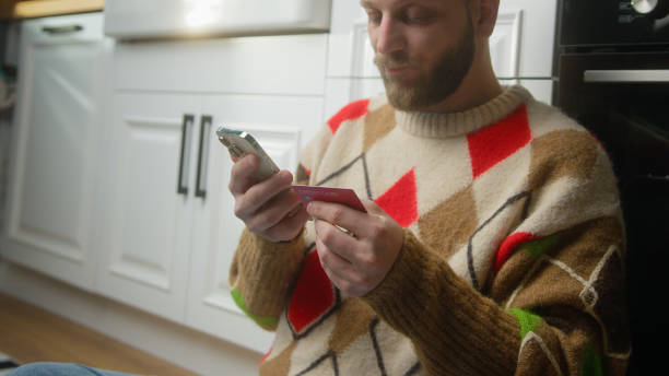 Adult man holding credit card and smartphone and due insufficient funds experiencing problems during payment online with credit card while sitting on the floor in kitchen Adult man holding credit card and smartphone and due insufficient funds experiencing problems during payment online with credit card while sitting on the floor in kitchen insufficient funds stock pictures, royalty-free photos & images