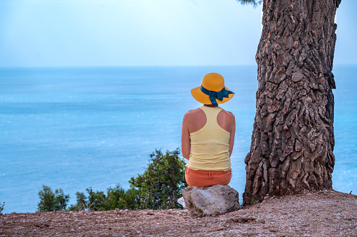 Tranquill scenery - one young beautiful woman with a hat seats at the cliff over the Ionian sea
