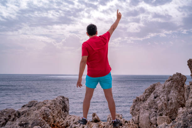 un joven con una camiseta roja se para al borde de un acantilado celebrando el logro - god freedom arms raised high angle view fotografías e imágenes de stock