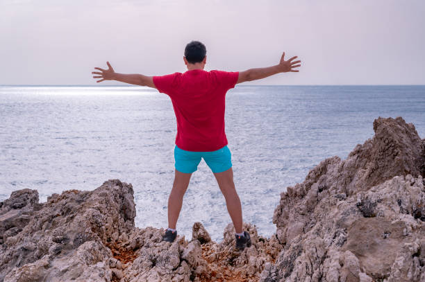 junger mann in rotem t-shirt steht am rande einer klippe und feiert erfolge - god freedom arms raised high angle view stock-fotos und bilder