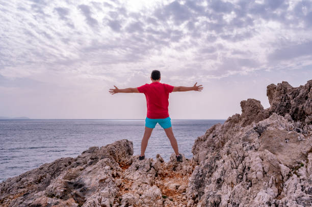 young man in a red t-shirt stands at edge of a cliff celebrating achievement - god freedom arms raised high angle view imagens e fotografias de stock