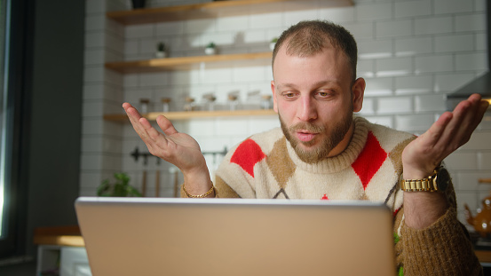 Shocked adult man sit in kitchen surprised, stunned while watching movie or video or reading news on laptop at home