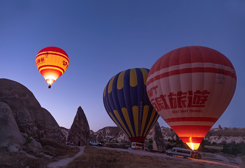 a hot air balloon full of tourists, about to take off at dawn between fairy chimneys, service vans around the balloons and other balloons ready to take off, one balloon in the air with flames in the burner, horizontal