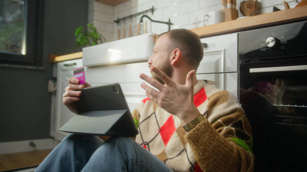 Sad man holding credit card and tablet and due insufficient funds experiencing problems during payment online with credit card while sitting on the floor in kitchen Sad man holding credit card and tablet and due insufficient funds experiencing problems during payment online with credit card while sitting on the floor in kitchen insufficient funds stock pictures, royalty-free photos & images