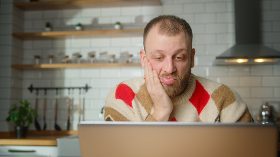 adult man sit in kitchen surprised, stunned while watching movie or video or reading news on laptop at home