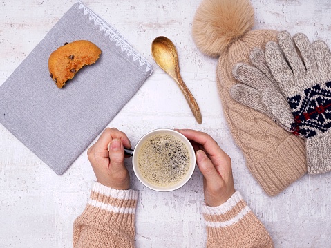 A flatlay photo of a woman's hands holding a cup of coffee, a wooden spoon, cookie, gloves, and a hat on the side.