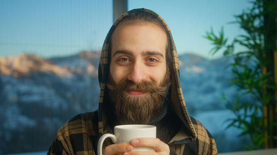 Portrait of bearded young man wearing hoodie drinking hot coffee or tea looking at camera smiling while sitting on couch in living room