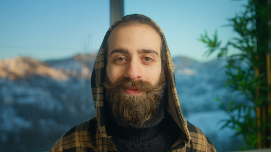 Portrait shot of bearded young man wearing hoodie looking at camera and smiling with snowy mountain background