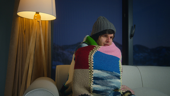 Young man wear scarf and beanie covering blanket himself for cold while sitting on couch in living room. Man expresses a sense of sadness and discomfort in the cold at home
