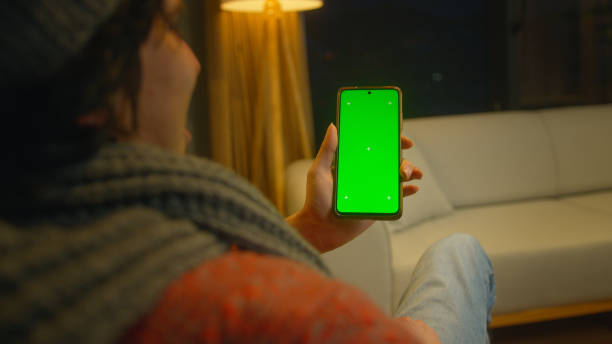 istock smiling man using, tapping on screen smartphone with green mock-up screen. Male sitting on sofa, relaxing at home. Close-up over the shoulder shot 1973659983
