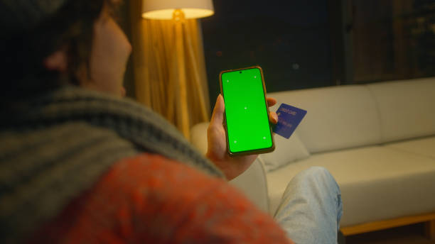 istock Man holding credit card, using, tapping on screen smartphone with green mock-up screen. Close-up over the shoulder shot 1973660492