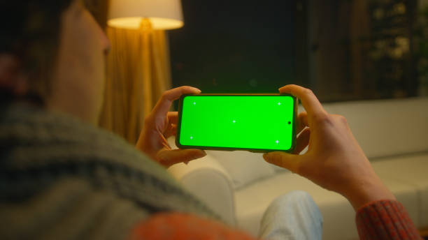 istock Young man watching video on his smartphone with green screen mock up display, which he holds horizontally. Male sitting on sofa relaxing at home. Close-up over the shoulder shot 1973661010