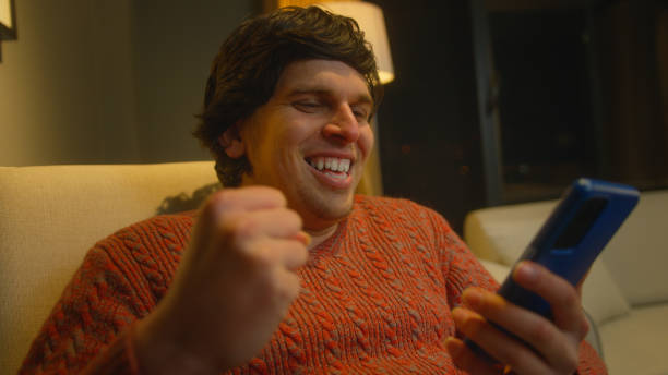 istock oung man sitting on couch using smartphone check message read notice receive good news on e-mail celebrate great opportunity feels happy in living room at night 1973664670