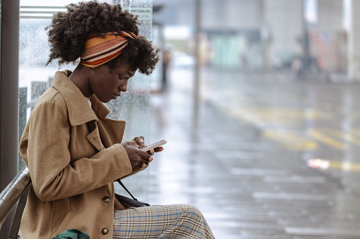 Side view shot of young African American woman at the bus stop. She is waiting for a bus and using smart phone in the city on a rainy day