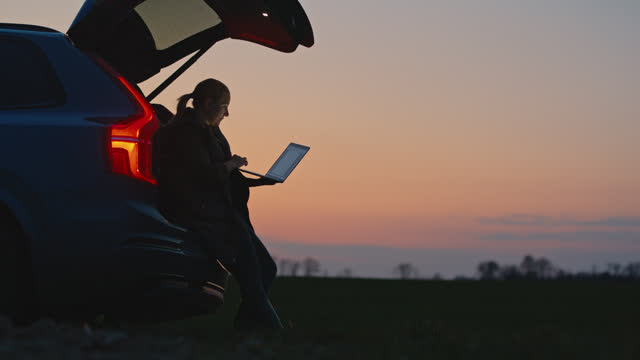 SLO MO Lockdown shot of Expectant Woman Using Laptop While Leaning on Car Trunk in Farm during Sunset. Female Farmer Holding Digital Tablet in Farm Field.