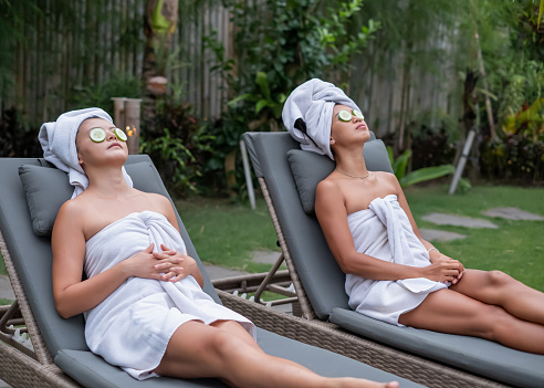 Side view shot of two Asian women lying on the tanning bed with refreshing cucumber slices on their eyes. They're both wrapped in towels while resting in luxury spa resort garden
