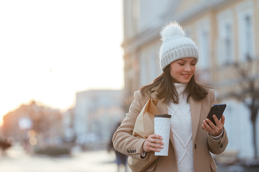 A young Caucasian woman strolls down the street, her attention captured by the smartphone in her hand. In her other hand, she clutches a reusable coffee cup. Dressed for the winter season, she is cozily wrapped in a beige coat, complemented by a white sweater and topped with a snug white cap.