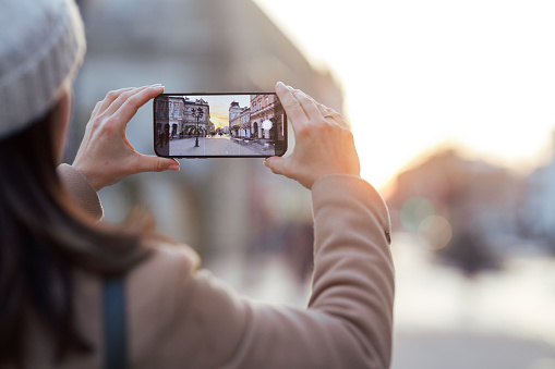 A Caucasian woman, clad in a stylish beige coat and a snug white cap, captures the breathtaking moment of a winter sunset in the city, immortalizing the picturesque scene with her smartphone camera.