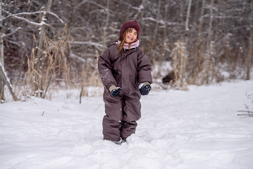 A sweet little girl is seen walking in the woods as she as she enjoys the cool fresh air and the crunch of fresh snow beneath her feet.  She is dressed warmly in a snowsuit, boots, hat and mittens.