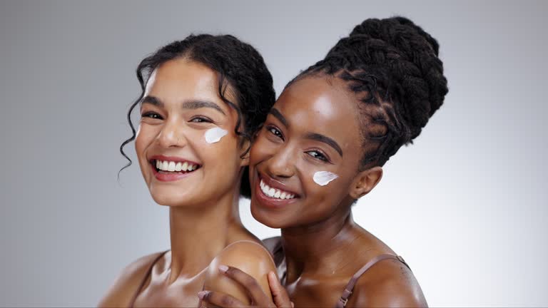 Women, cream and beauty with diversity in studio with facial treatment, happiness or glowing skin. Friends, model and portrait with smile for cosmetics, dermatology or body care on gray background