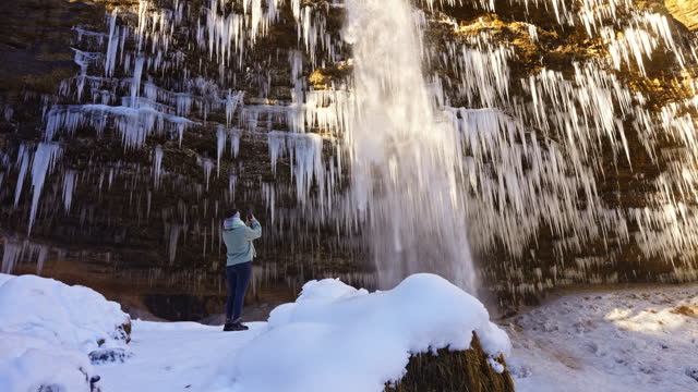 SLO MO Female Hiker on Snowy Rocks Photographing Peričnik Falls with Icicles Covered Cliff in Triglav National Park, Mojstrana