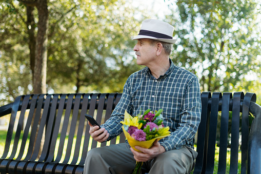 Anxiously expectant, the elderly man sits on the park bench, bouquet in hand, his heart brimming with anticipation as he eagerly awaits the arrival of his beloved partner for a rendezvous amidst nature's tranquility.