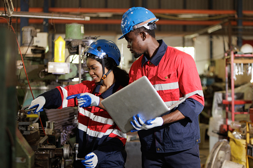 Two engineer heavy Industry Engineers Wearing Safety Uniform and Hard Hats Working on Laptop Computer. African American Technician and Female Worker Talking on a Meeting in a Factory.