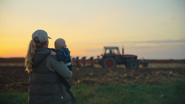 SLO MO Woman Farmer Carrying Baby Boy and Watching Tractor Plowing Field against Sky During Sunset. A Farmer and Her Son in the Field.