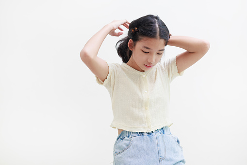 Portrait of a pretty and attractive little Asian girl is captured looking down while tying her hair, creating a charming image set against a white background.