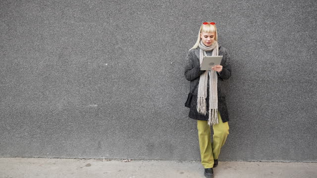 Happy hippie girl browsing a tablet working freelance and leaning on a wall of office building in the street. Hipster woman online work while waiting colleagues for staff meeting
