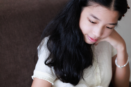 A carefree and relaxed young Asian girl sitting on a sofa with her smartphone. She's scrolling through social media, browsing the internet, shopping online, or chatting with friends, capturing a moment of leisure and digital connectivity.