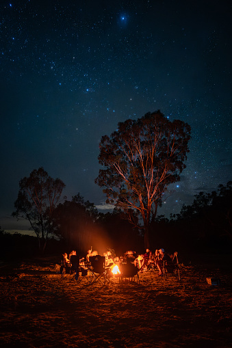 A group of people camping in the Australian outback, around a campfire with the stars shining above them.