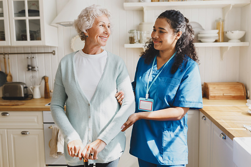 Medical assistance at home. Pretty african american female volunteer in blue uniform helping senior caucasian woman walk with walking stick, holding her by hand, looking at each other with smile