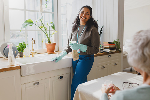 Selective focus on happy african american volunteer helping senior lady to clean house, standing in kitchen in raisin gloves washing sink with detergent, talking to elderly female sitting at table