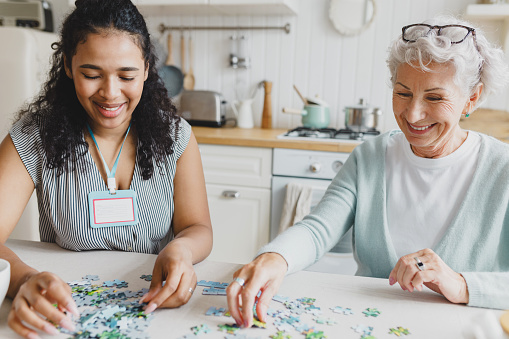 Portrait of two people at kitchen table, african american female volunteer spending leisure time with senior lady, playing together puzzle game, chatting and having fun. Social support