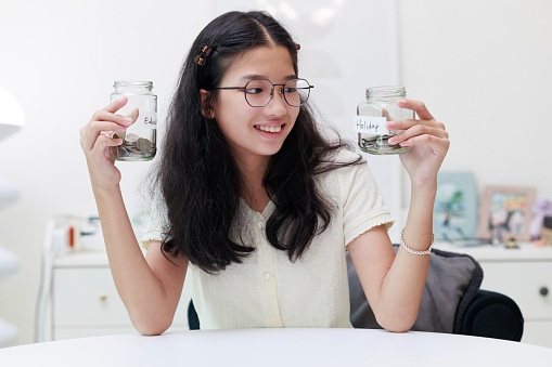 A young Asian girl is captured putting coins into a jar, symbolizing her commitment to saving money for both education and future holidays.