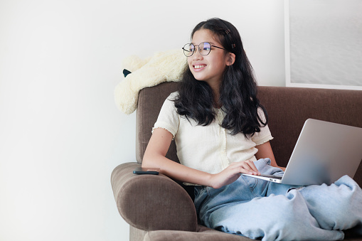 A carefree and relaxed young Asian girl sitting on a sofa with her laptop. She's looking away while scrolling through social media, browsing the internet, shopping online, or chatting with friends, capturing a moment of leisure and digital connectivity.