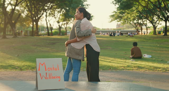 Two young Asian women sharing a supportive embrace in a public park during a mental health awareness event, highlighting compassion and care. Outdoor mental wellbeing community support.