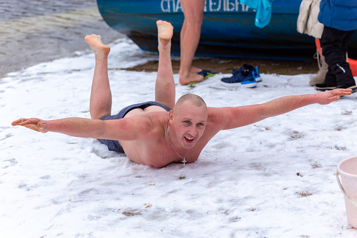 Grodno, Belarus - January 28, 2024: A happy man in his underpants lies in the snow on the riverbank during the traditional annual Temper-fest race. A scuba diver lifeguard is on duty in the water for safety.