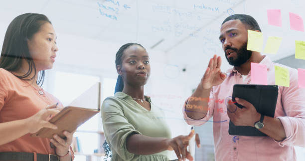 istock Business people, teamwork and meeting for schedule planning, sticky notes or strategy on glass board at office. Group of employees in team brainstorming, ideas or reminder for agenda together 1974544061